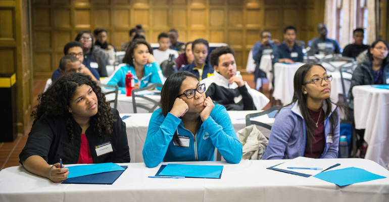 People seated at round tables with blue folders during a conference or workshop, listening and taking notes in a large room with rows of attendees behind them.