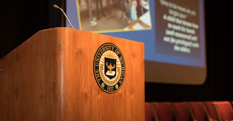 Wooden podium with a black-and-gold university seal, a microphone stand rising from the left, and blurred event posters and red auditorium seats in the background.