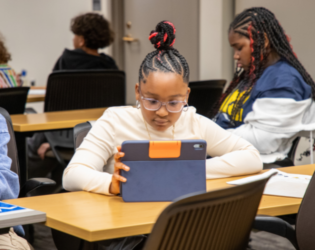 A student with a high bun and glasses sits at a classroom desk, holding a tablet with an orange case while studying; other students sit nearby in a lecture room.