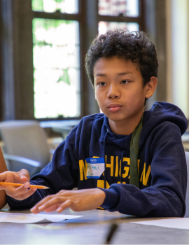 A young person in a dark hoodie sits at a classroom table, looking toward the camera while working with hands near papers; another student sits nearby, with windows and desks in the softly lit background.