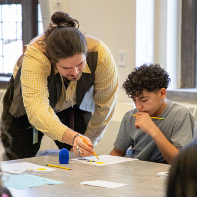 An adult and a child sit at a table using colored markers and papers while working on an art or craft project in a bright room with large windows.