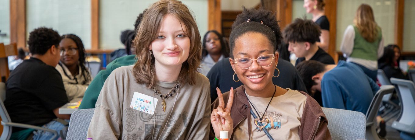 Two students sit at a table covered with colorful craft supplies, smiling at the camera during an indoor event, with other attendees in the background and a large blue banner overhead.