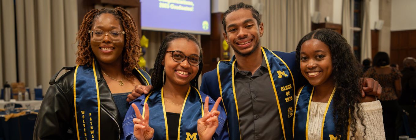 Four students in navy graduation gowns with gold trim pose together indoors, smiling and making peace signs, with a projector screen and banquet tables in the background.