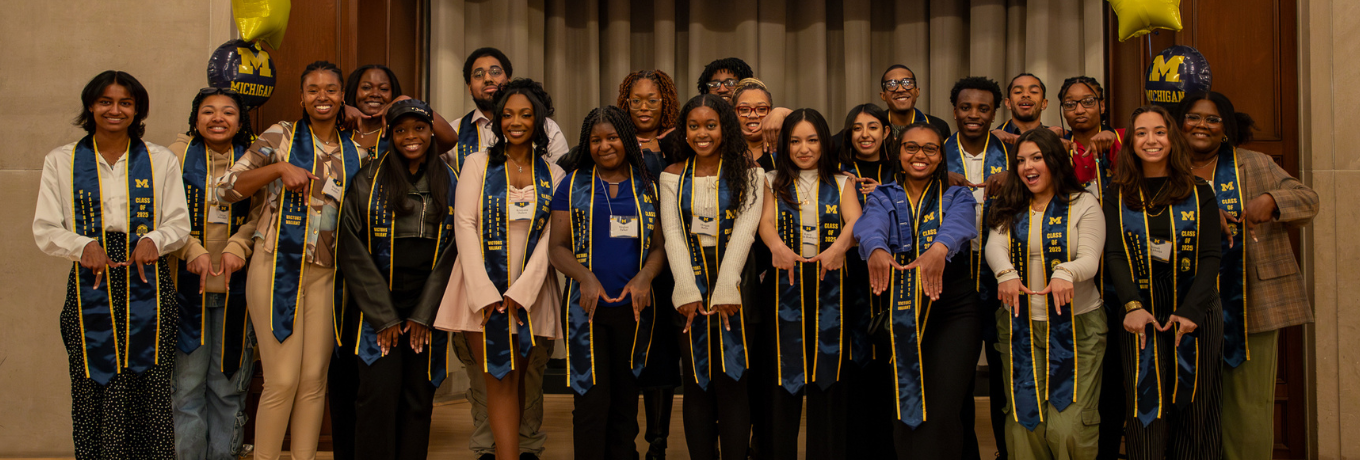 Group of graduates in academic gowns and stoles pose on a stage with blue and gold decorations, while a screen behind them reads “Congratulations, 2025 Graduates!”