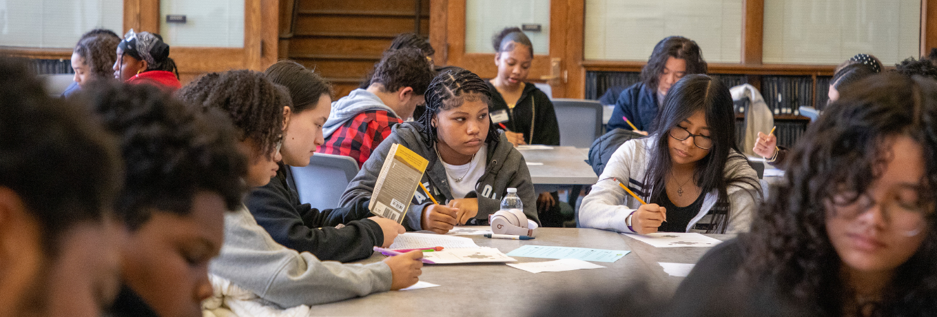 Students sit around a long table working on papers inside a school room.