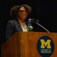 A speaker stands at a wooden podium with the University of Michigan logo, speaking into a microphone; a small flower arrangement sits on the podium, with another person seated to the right against a dark backdrop.