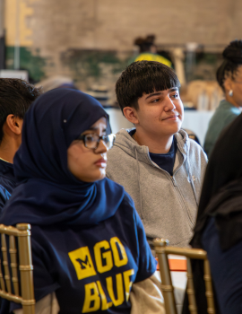 A group of students in a classroom sit at desks, wearing dark hoodies and headscarves, talking and looking toward the front as notebooks and papers rest on the table.