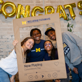 A group of people pose behind a large poster board on a brick wall, with “CONGRATS” in gold letters above; the board shows a “Now Playing” graphic and faces.