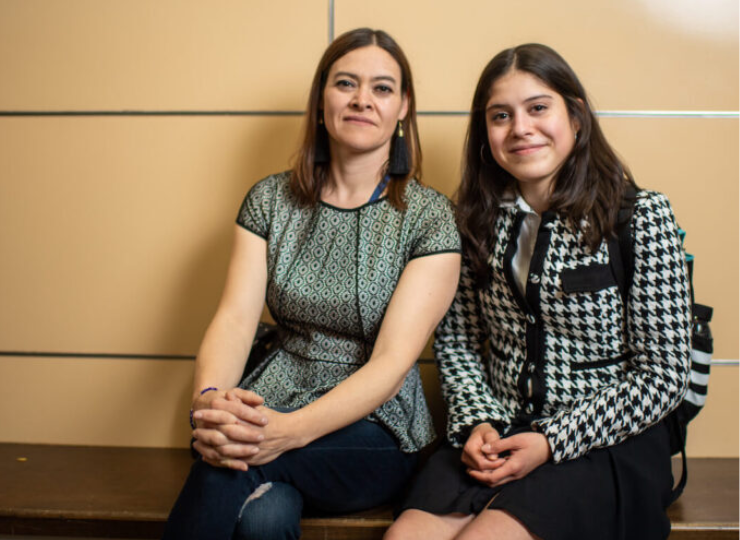 Two seated women posing for a photo against a beige wall with horizontal lines, one in a green patterned top and jeans, the other in a black-and-white houndstooth dress.