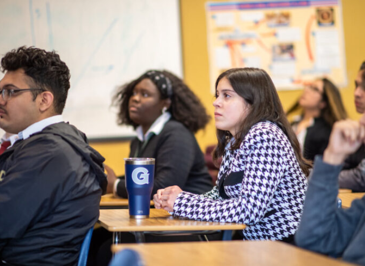 Students seated in a classroom, listening to a discussion; one person wears headphones, another holds a blue drink cup, and a whiteboard and yellow bulletin board are visible in the background.
