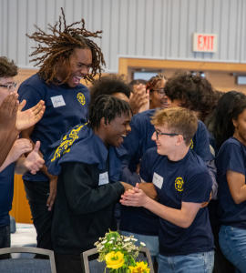 Students in blue shirts stand in a group indoors, smiling and clapping while looking toward a table with a green-and-yellow decorated centerpiece; an EXIT sign glows on the back wall.