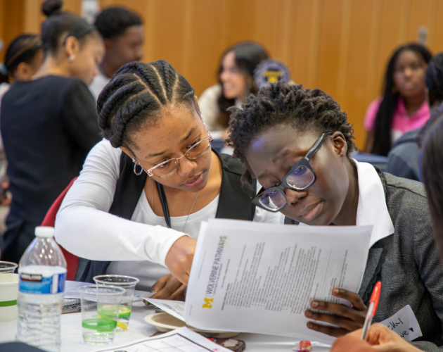 Students sit at classroom tables, reading worksheets and writing with pens; one student in front wears glasses and smiles while studying a printed handout.