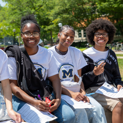 Four students in matching T-shirts sit on a bench outdoors in a park, smiling and talking, with trees and a lamppost in the background.