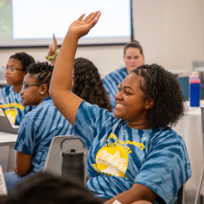 Students in a classroom wearing matching blue T-shirts sit at desks while one student raises a hand during a lesson, with a teacher standing near a projection screen in the background.