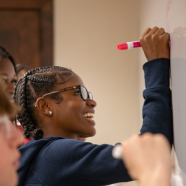A student wearing glasses smiles while writing with a pink marker on a whiteboard, with other students seated nearby and a classroom wall in the background.