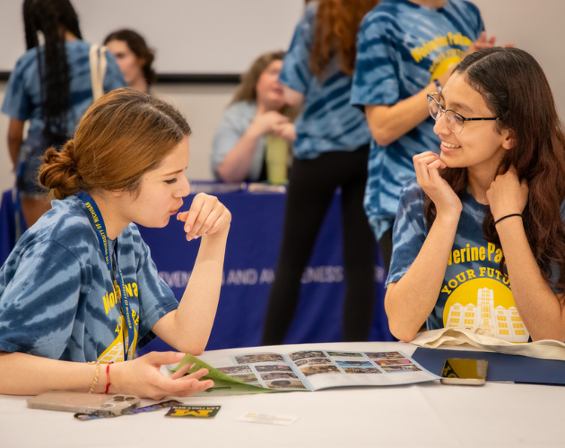 Two students in blue tie-dye shirts sit at a table looking at a photo booklet and smiling while talking, with other students blurred in the background during a classroom activity.