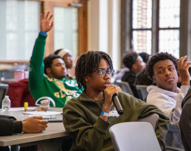 In a classroom, a student in a green hoodie speaks into a microphone while others sit at desks and raise hands; adults and windows fill the background.