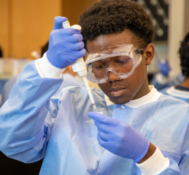 A person in a lab coat and blue gloves adjusts a face covering while wearing safety goggles and applying white cream to the forehead; others work in the background in a clinical setting.