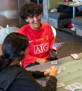 A group of people sit around a table playing a card game indoors, with several cards spread on a green felt surface and one person in a red shirt smiling while another leans in to play.