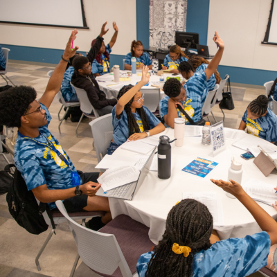 A group of students in blue shirts sit around classroom tables with notebooks and cups, raising their hands during an activity, while large projection screens hang on the walls.