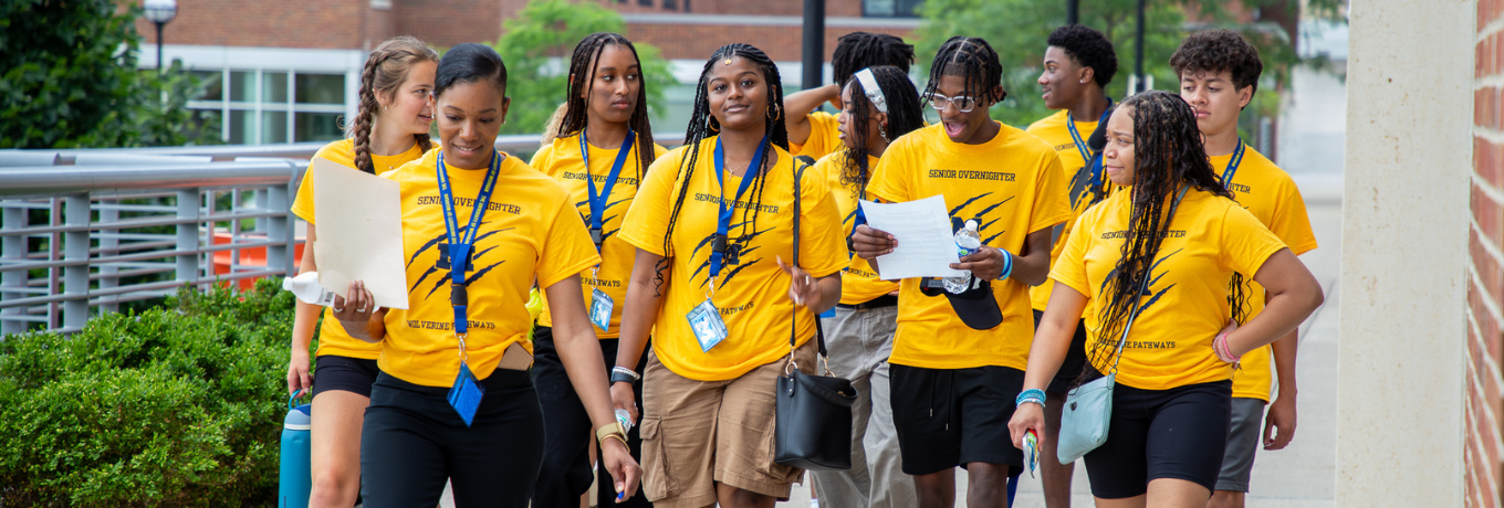 Students in matching yellow T-shirts with lanyards walk together down a campus walkway, holding papers and backpacks, with brick buildings and hedges in the background.