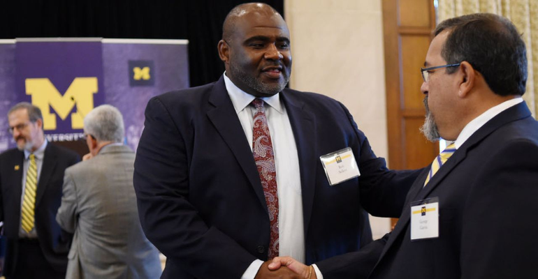 Two men in suits greet and shake hands at a conference, wearing name badges; a purple event banner with a large “M” and “M” logo is visible in the background.