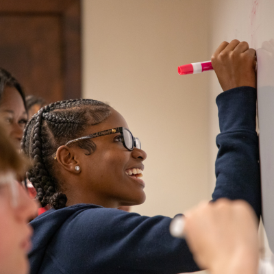 Students in a classroom smile while writing on a whiteboard with markers, with one person reaching up to add notes and others watching from the foreground.