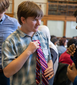 Students in a school setting stand close together as one adjusts another’s red, white, and blue striped tie while others watch and smile.