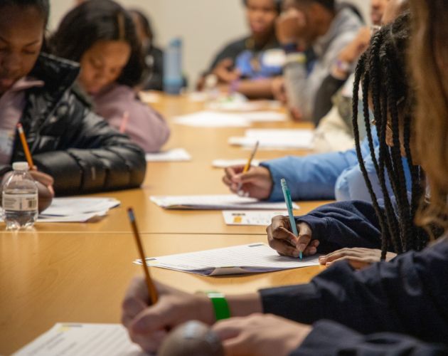 Students seated at desks in a classroom, writing with pencils on worksheets while others work in the background.