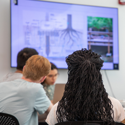 A person with curly dark hair sits at a desk in a classroom or office, facing colleagues and a wall monitor displaying technical diagrams and a green image.