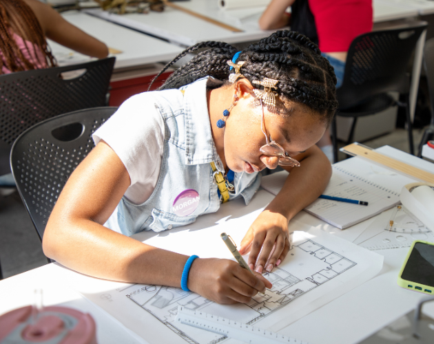 A student in a classroom leans over a large sheet of printed diagrams, drawing and writing with a pencil while working at a desk with other supplies nearby.