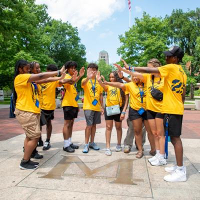A group of students in matching yellow shirts line up on a sunny outdoor walkway, facing away and high-fiving with arms out while standing near trees and a tall building in the background.