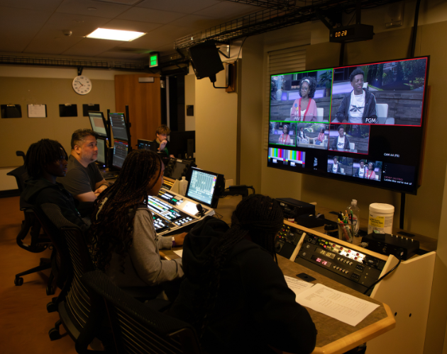 Recording studio control room with multiple people seated at mixing consoles and computers, while a wall-mounted monitor shows a live feed with several video windows and audio meters.