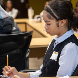 Student sitting at a desk in a classroom, writing with a pencil while other students sit in the background, with chairs and papers visible.