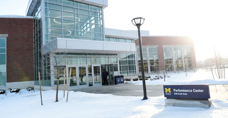 Modern brick and glass building with a central entrance, snow-covered ground, and a sign in the foreground reading “Performance Center.”