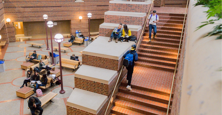 People gather on steps and platforms in a brick courtyard, with a few seated near benches and lamp posts under warm lighting.