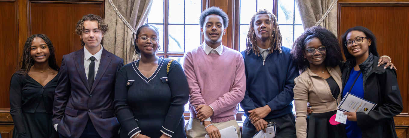 A group of seven students in formal clothes pose indoors in front of large wooden doors and tall windows, holding notebooks or folders and smiling at the camera.