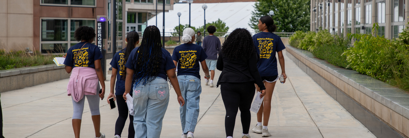 People walking down a campus walkway between modern buildings, with trees and shrubs along the sides under a partly cloudy sky.