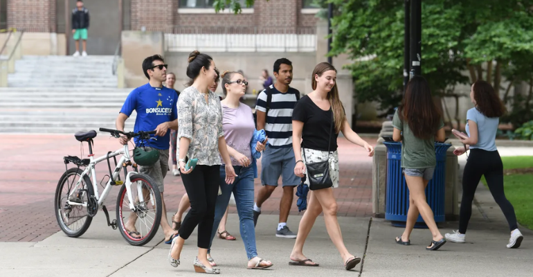 A group of students walks along a campus walkway with bicycles parked nearby, passing a brick building, trees, and a blue trash bin.