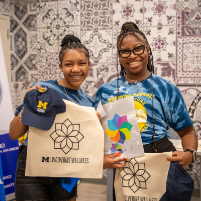 Two students in blue T-shirts stand side by side holding tote bags with printed line-art flowers at an indoor event booth with patterned wallpaper and Phi Beta Sigma banners.